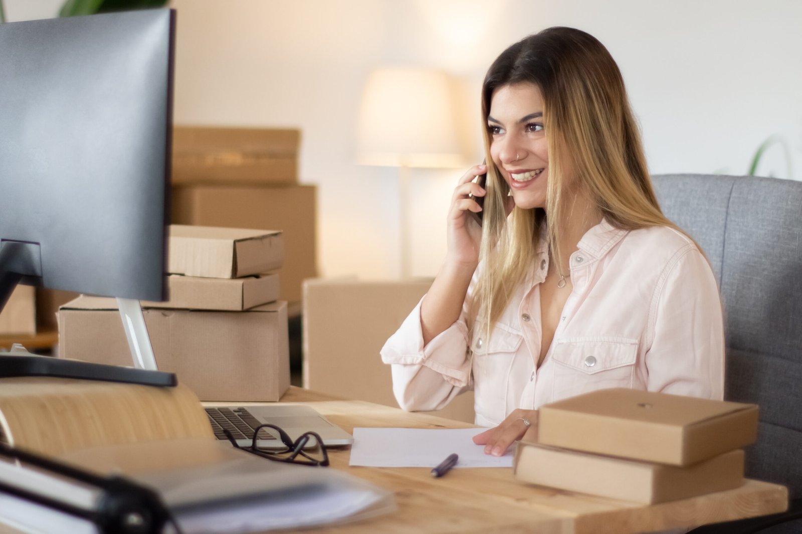 Young Online Shop Assistant Consulting Client Phone Looking Computer Screen Smiling Beautiful Caucasian Woman Working Home Office Surrounded By Cardboard Parcels E Commerce Concept 1 Scaled